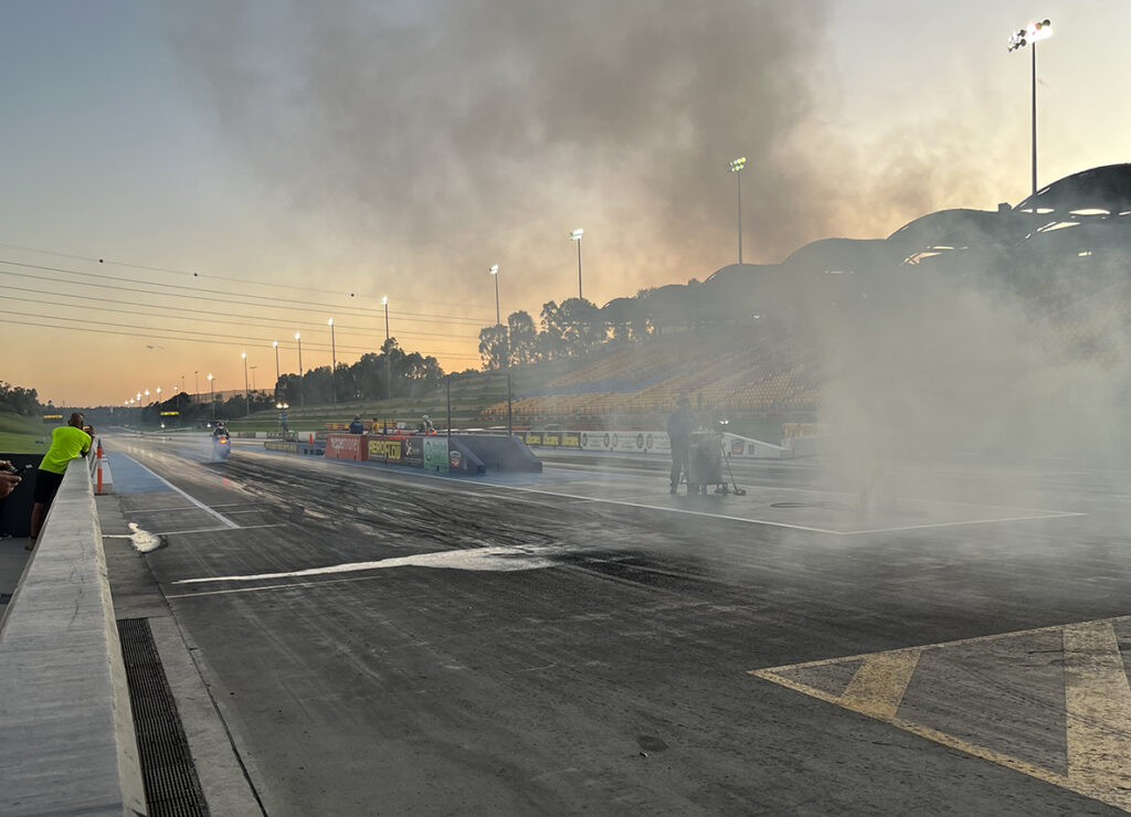 A motorcycle racer lines up at the start line of the quarter mile at Sydney Dragway on Bike Night after completing a burnout. The smoke from the burnout billows into the sunset sky. The tail lights of a motorcycle shine in the background as the racer waits for the green light to go.