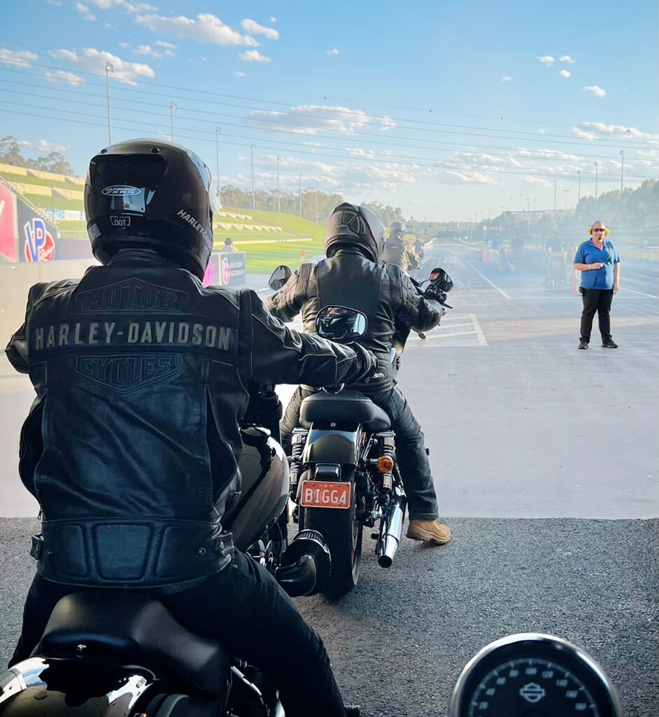 Motorcycle racers line up behind the start line of the quarter mile at Sydney Dragway on Bike Night. The picture is taken from a racer behind other motorcycle racers, looking down the barrel of the race track past their right shoulders. The sun casts deep shadows over the race track, the grandstand on the left glowing in the sunlight with a soft blue sky.