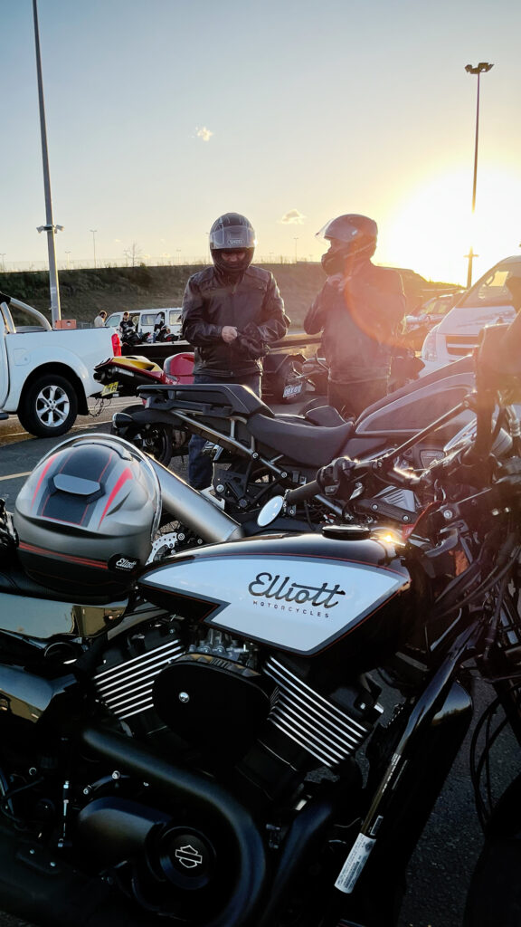 The sun sets on the horizon of a clear sky day while motorcycle racers put on on their helmets and gloves with their motorcycles. The sun reflects off the tank of a custom Elliott Motorcycles hooligan motorcycle in the foreground.