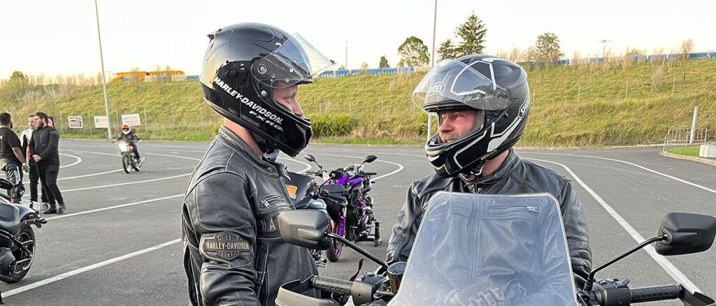 Elliott Andrews and Dan Lesnock talking at Sydney Dragway Bike Night. Dan sits on a motorcycle while Elliott stands beside him. Both wear motorcycle leathers and helmets.